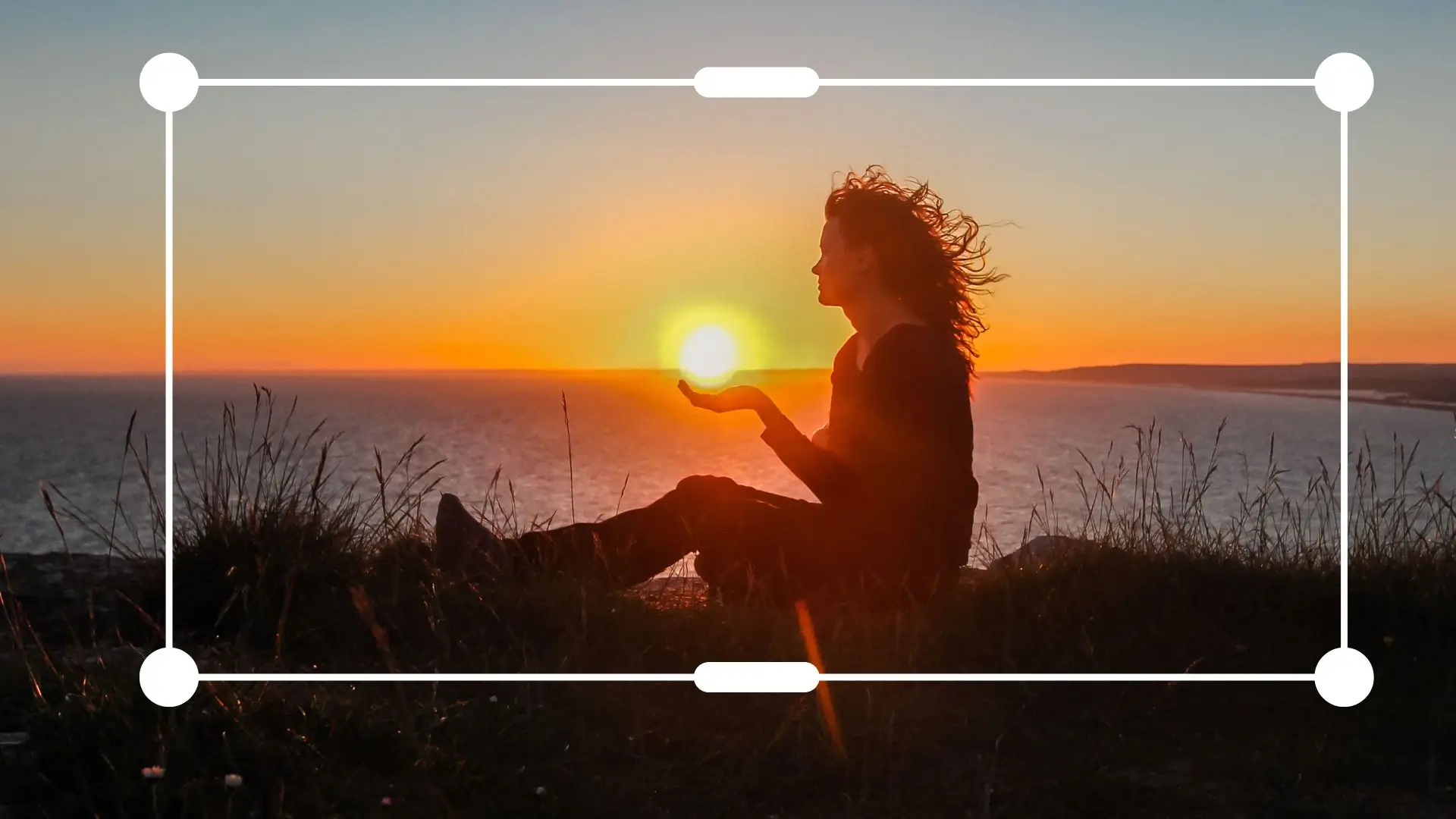 A woman sits on a hill, gazing at a vibrant sunset that paints the sky in warm hues.