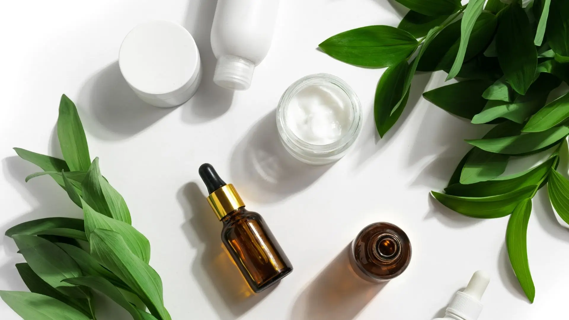 A collection of cosmetic products surrounded by green leaves on a clean white surface, showcasing natural beauty elements.‍