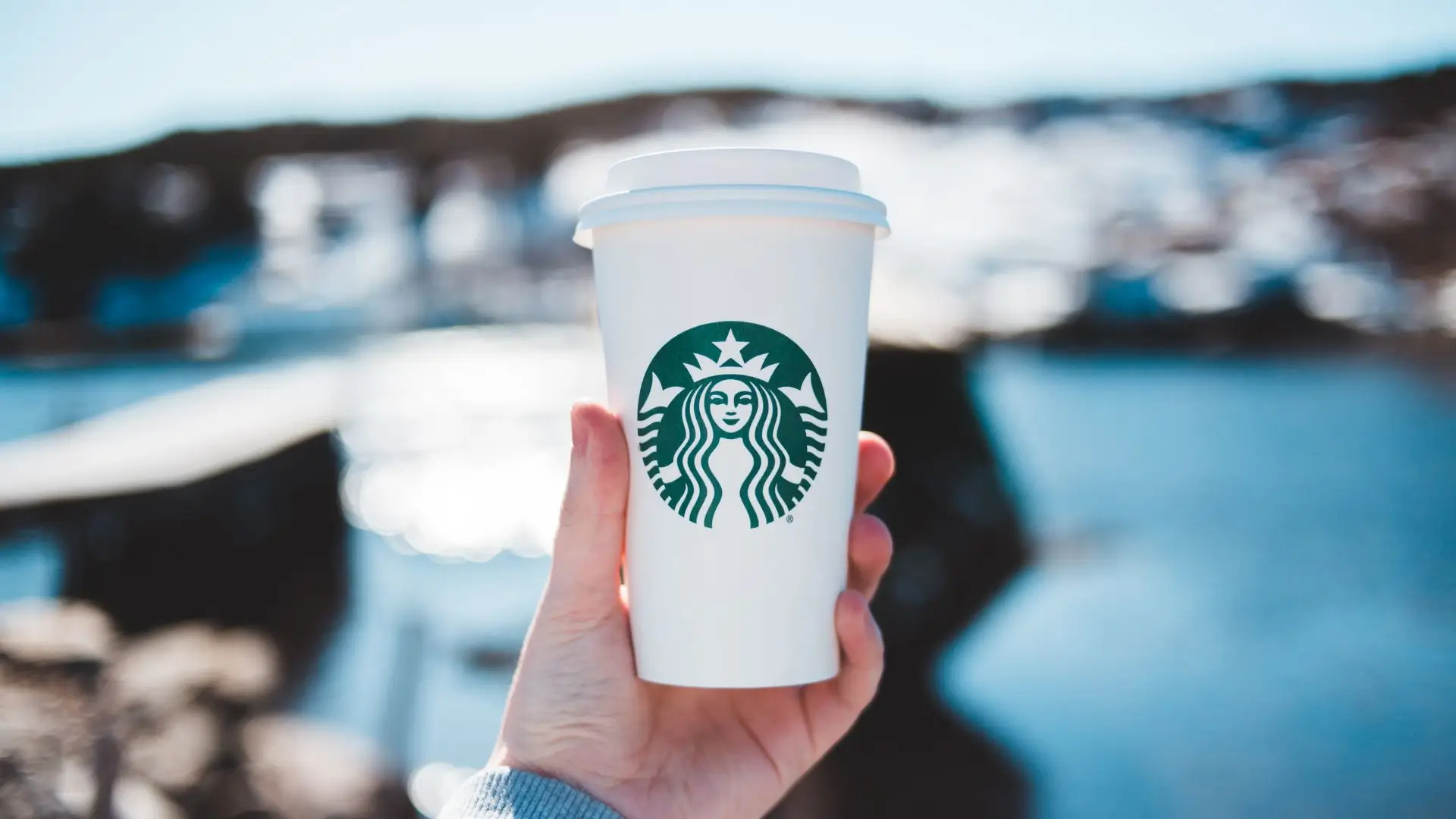 A person enjoys a Starbucks cup while standing by a serene lake, surrounded by nature's beauty.
