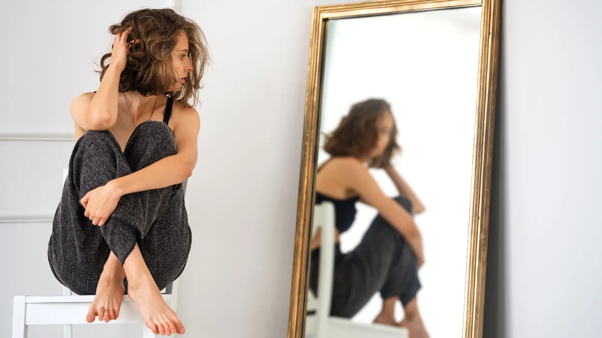 A woman sits in a chair, observing her reflection in the mirror with a contemplative expression.‍