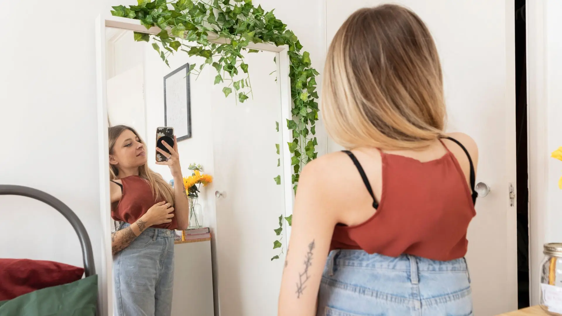 A woman in a red tank top and denim shorts smiles while taking a selfie in front of a mirror.