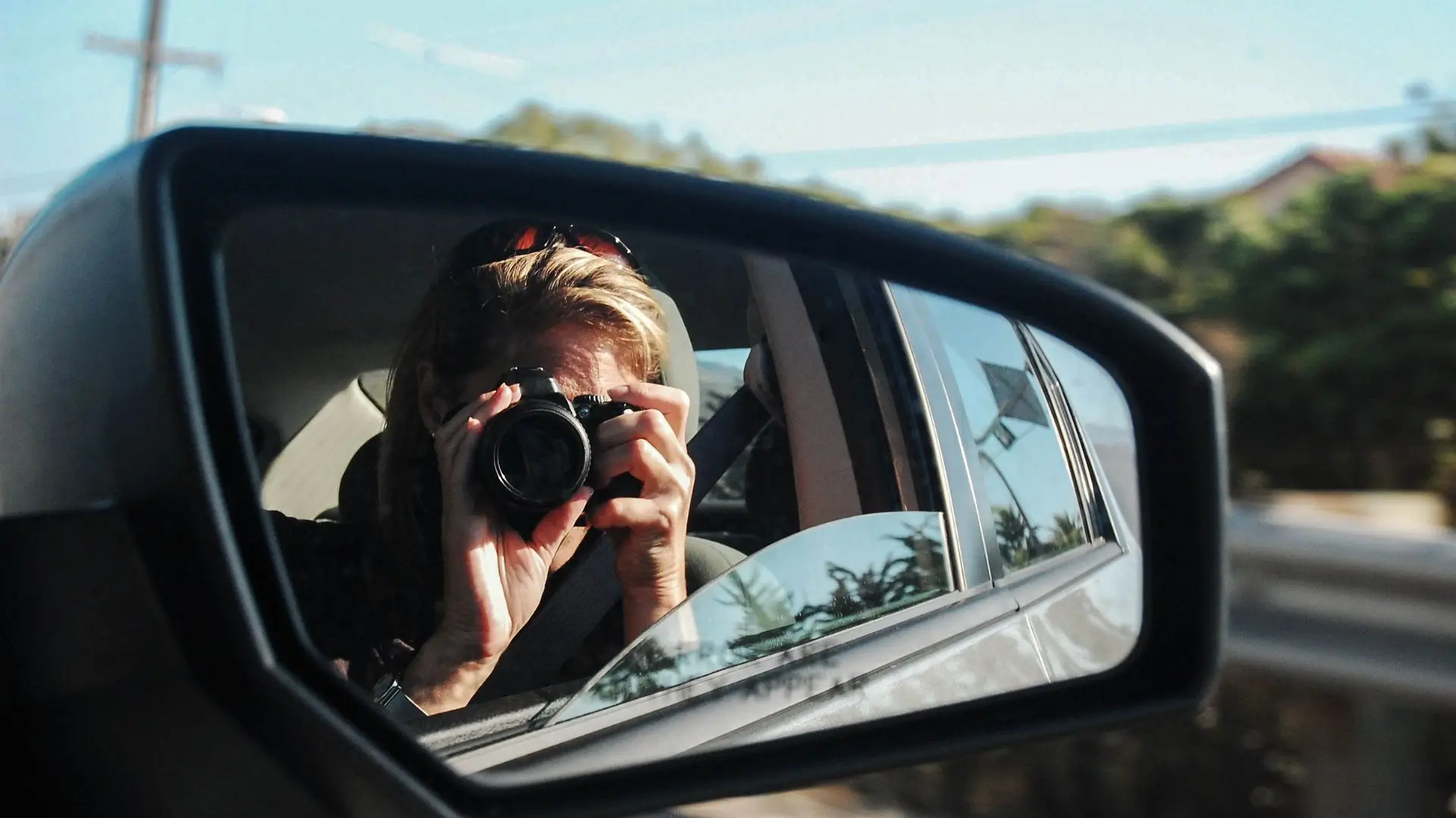 A woman captures a photo using the rear view mirror, reflecting her focused expression and the surrounding scenery.