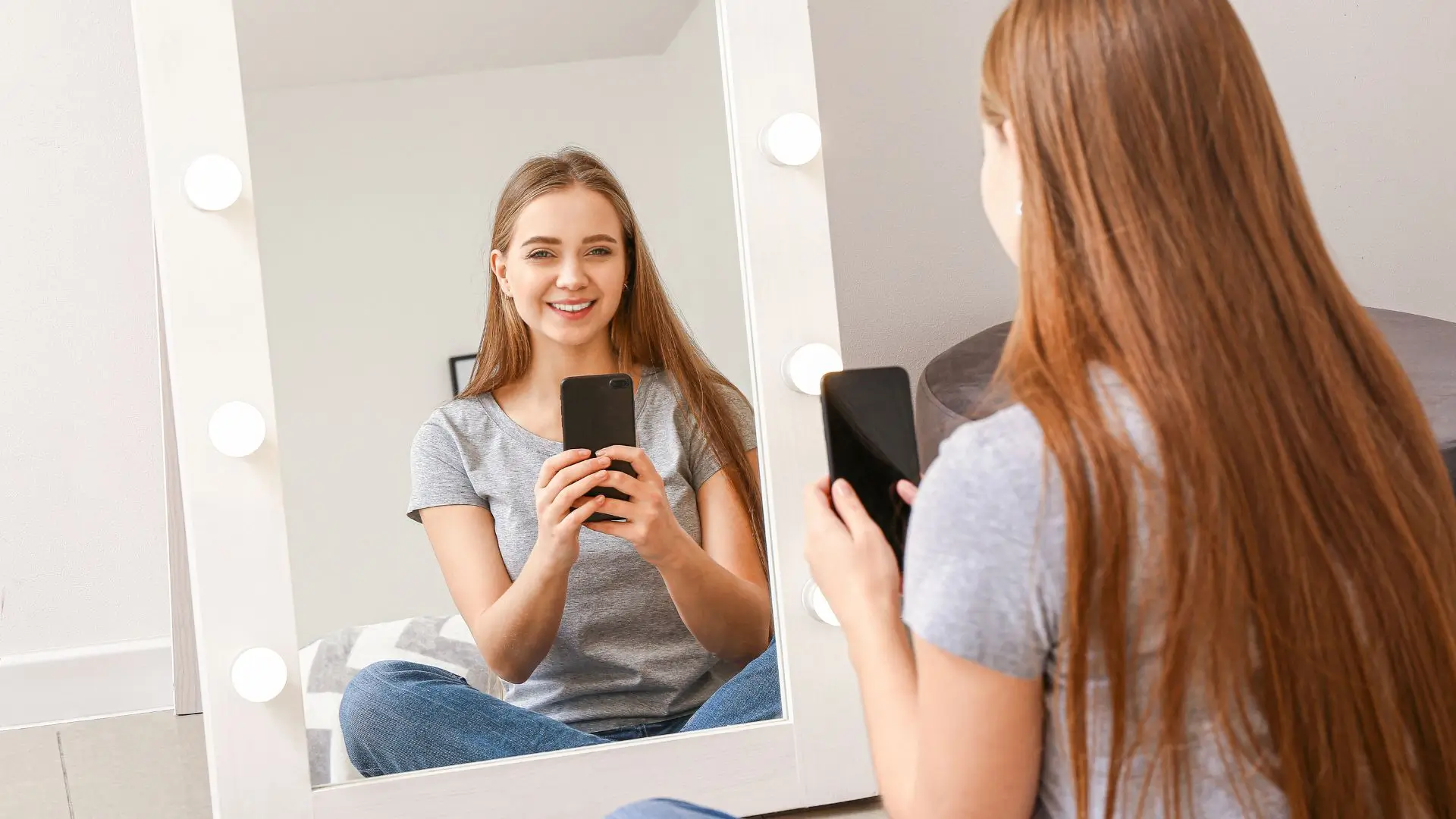 A woman sits in front of a mirror, holding her phone, reflecting on her appearance with a thoughtful expression.‍