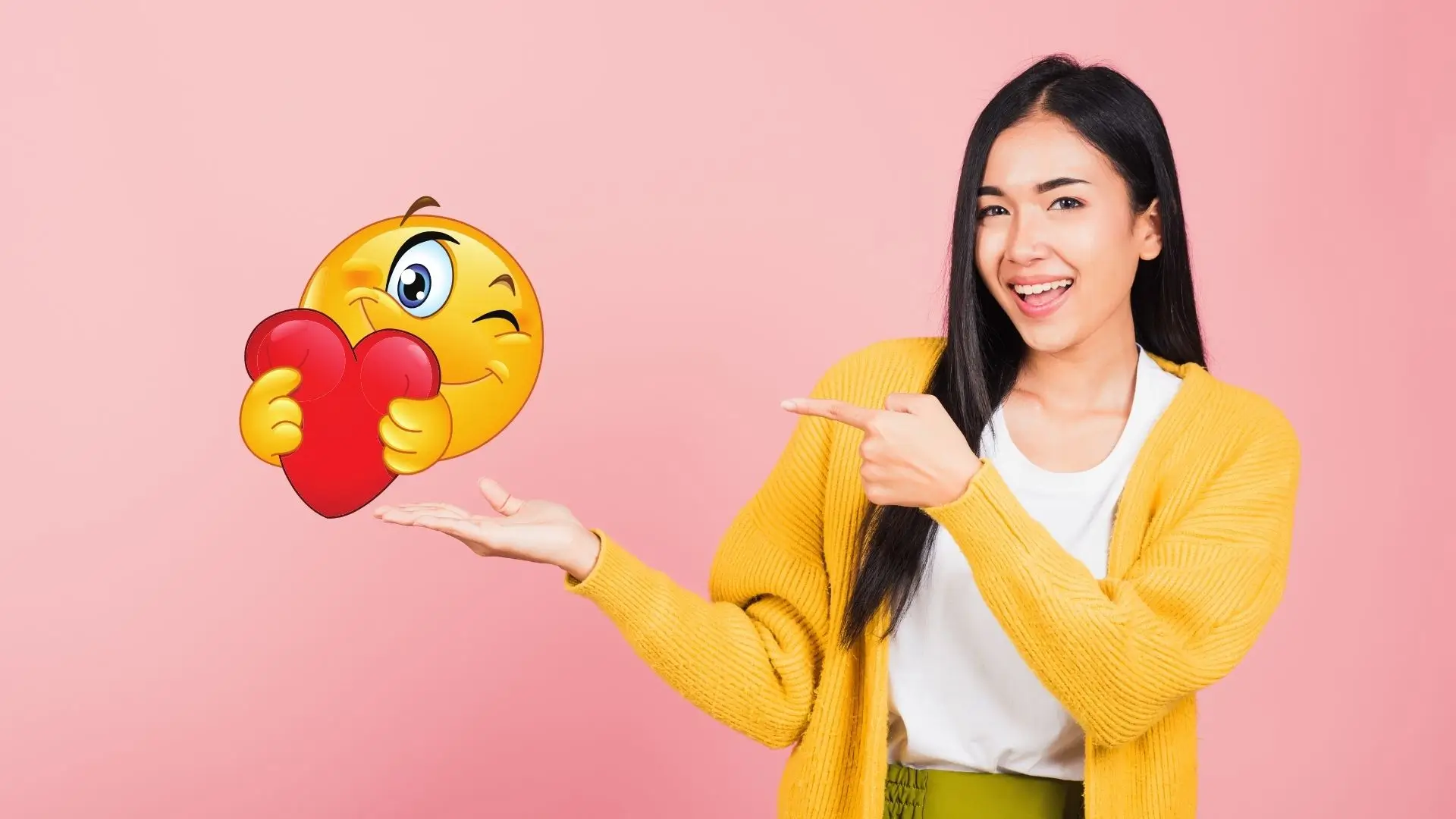A woman smiles while holding a heart emoticon against a soft pink background, conveying love and positivity.